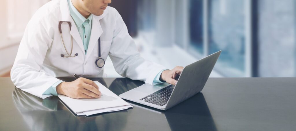 Male doctor working on desk with laptop computer and paperwork on his desk looking at a new ehr vendor