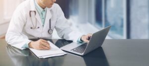 Male doctor working on desk with laptop computer and paperwork on his desk looking at a new ehr vendor